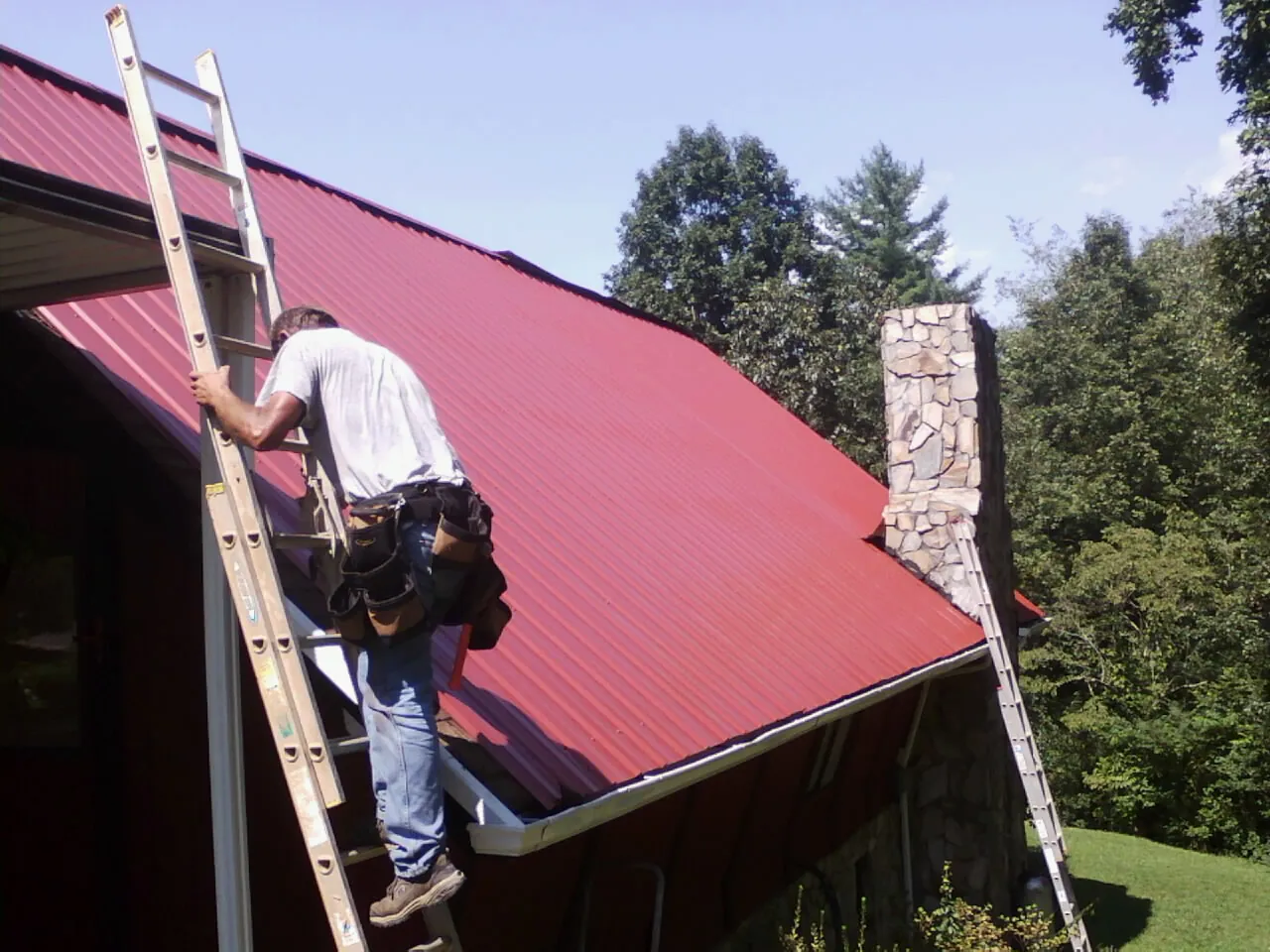 Roofer on a ladder beside a red metal roof and stone chimney — Soffit & Fascia Repair by Roofing Brothers in Lorton